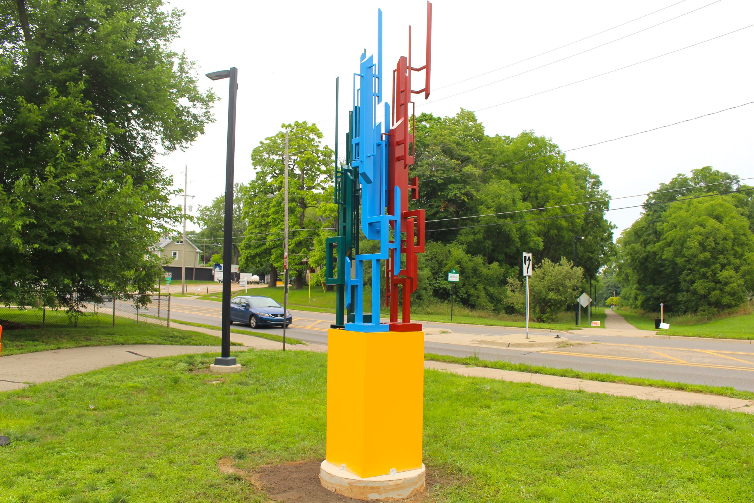 A multi-colored metal sculpture along a street and trail 
