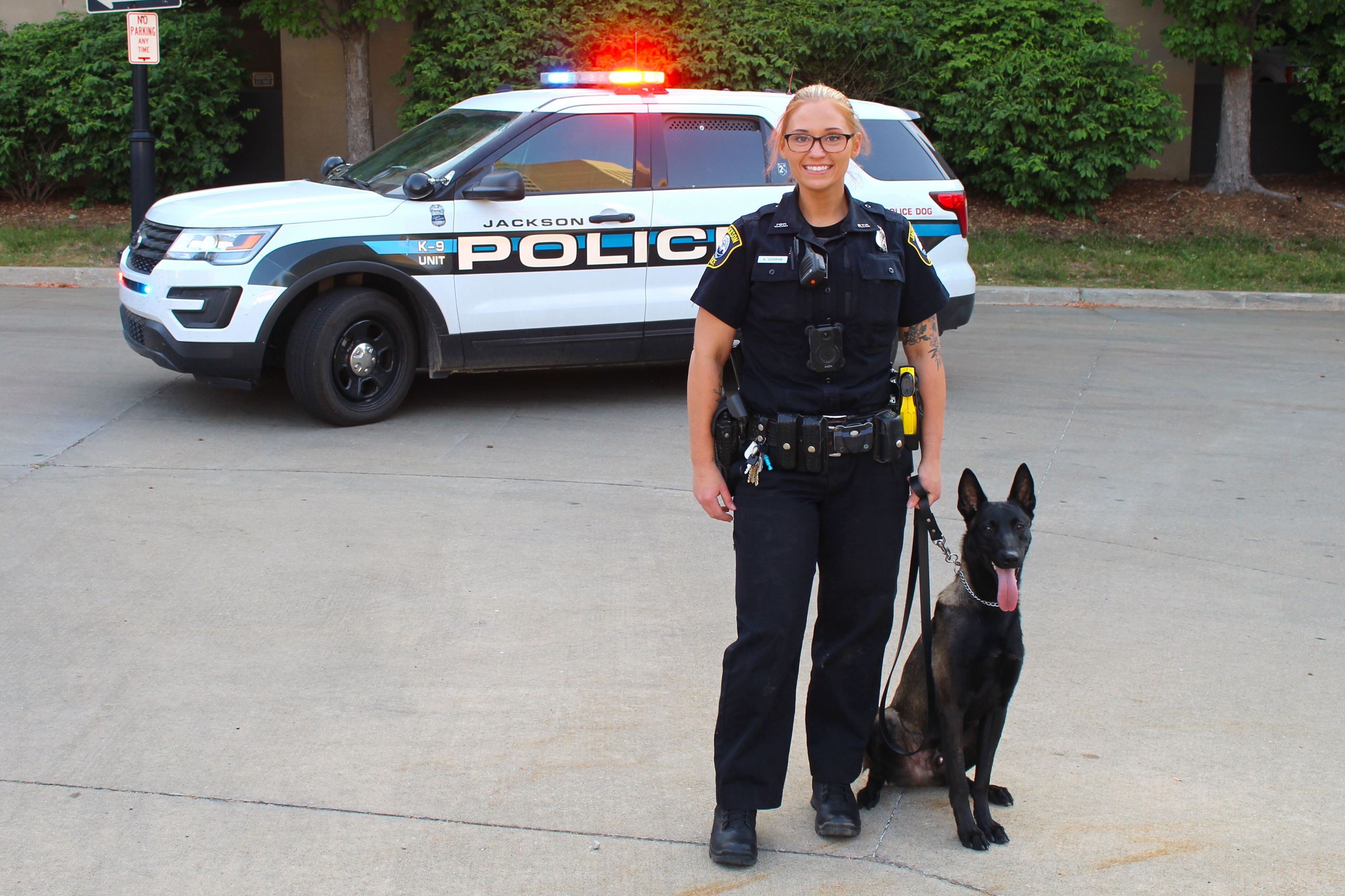 A young white woman stands in front of a police car smiling with a black dog sitting on a leash