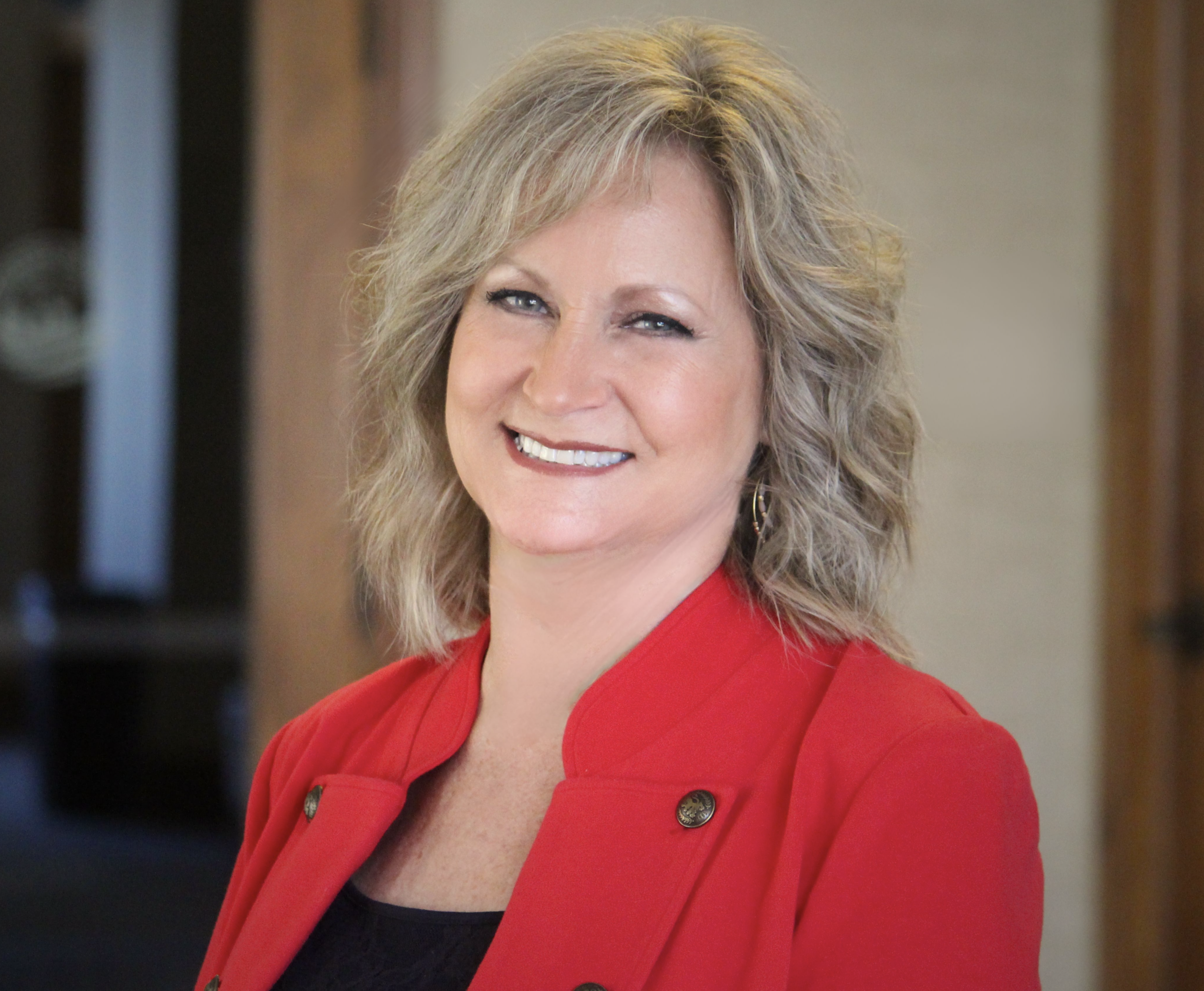 A white woman with blonde hair smiles wearing a red jacket 