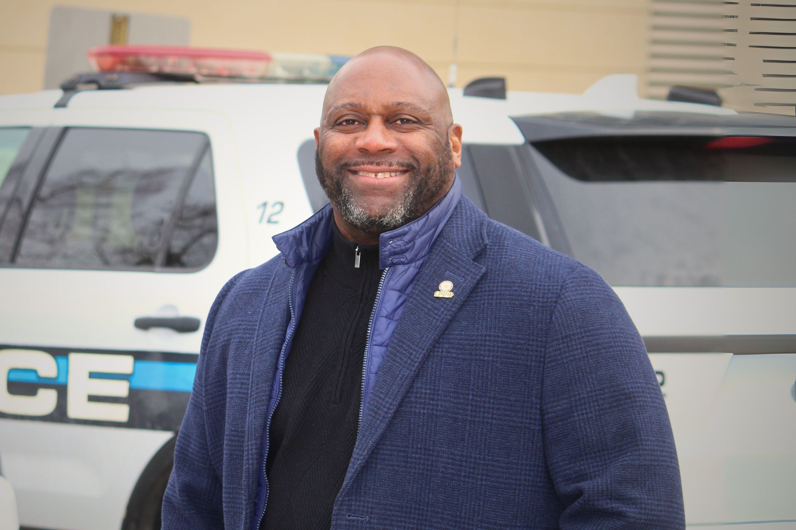 A man in coat smiles in front of a police car 